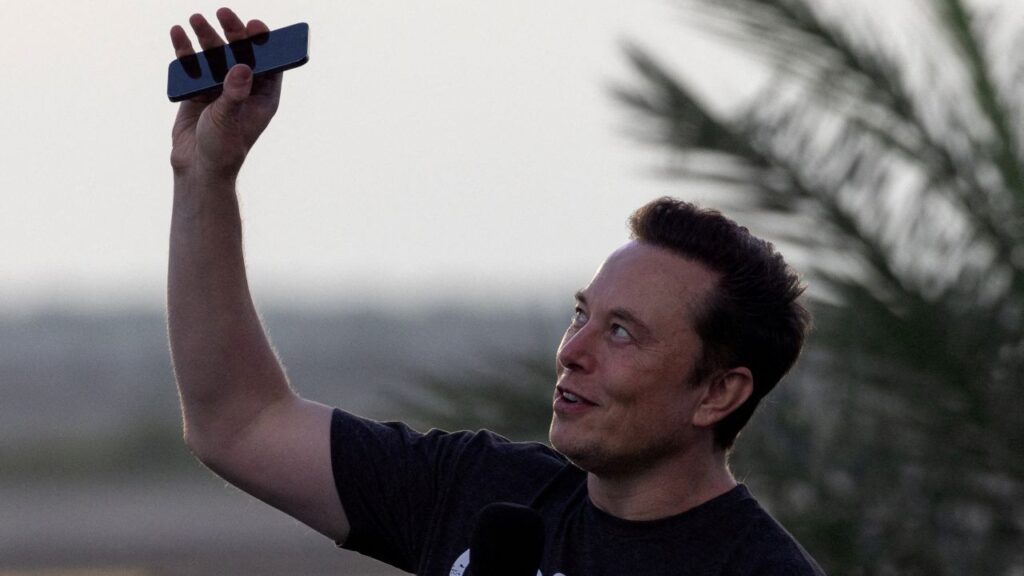 An image Elon Musk dressed in a dark t-shirt holding up a cell phone to the sky at dusk