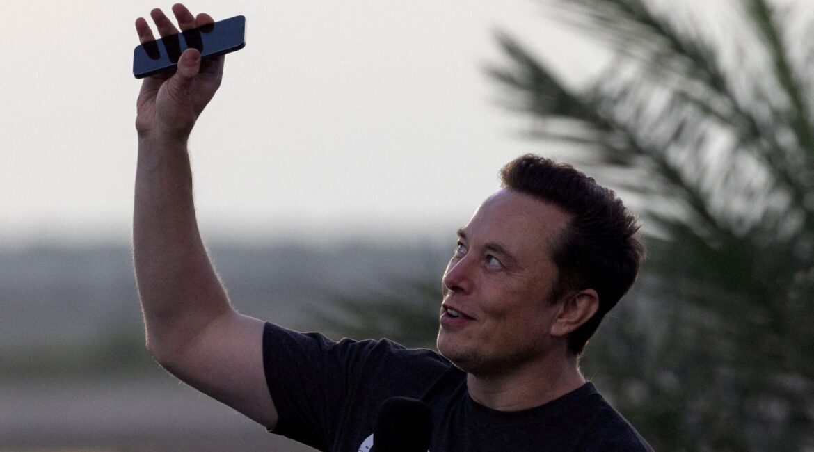 An image Elon Musk dressed in a dark t-shirt holding up a cell phone to the sky at dusk