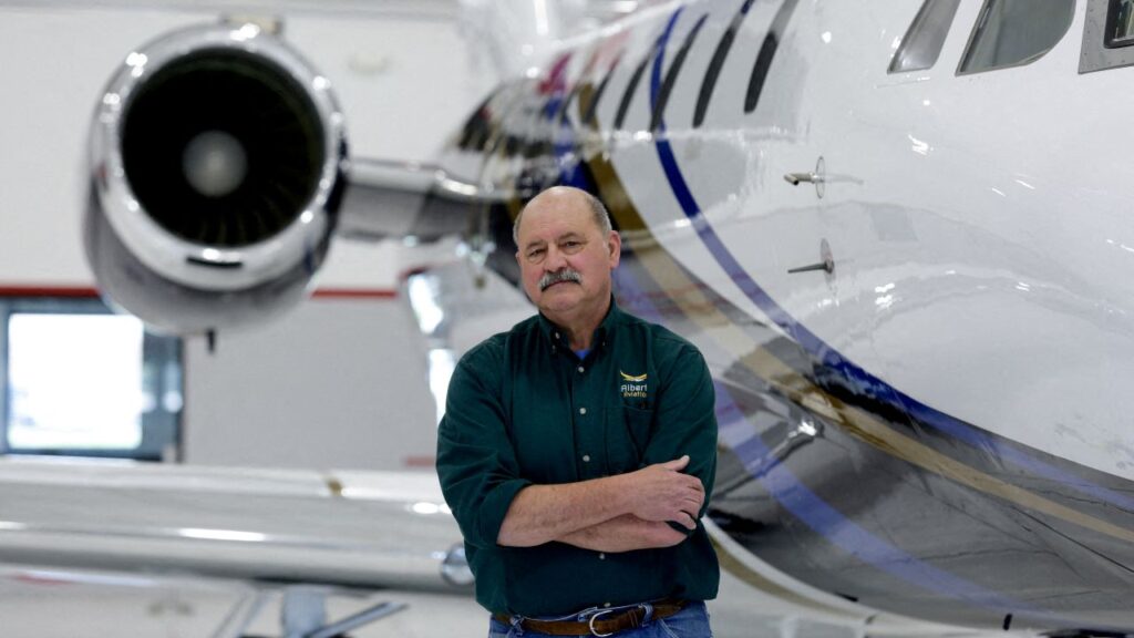 Image of a bald man with a mustache, dressed in a dark shirt and blue jeans, standing next to a jet airplane in a hangar