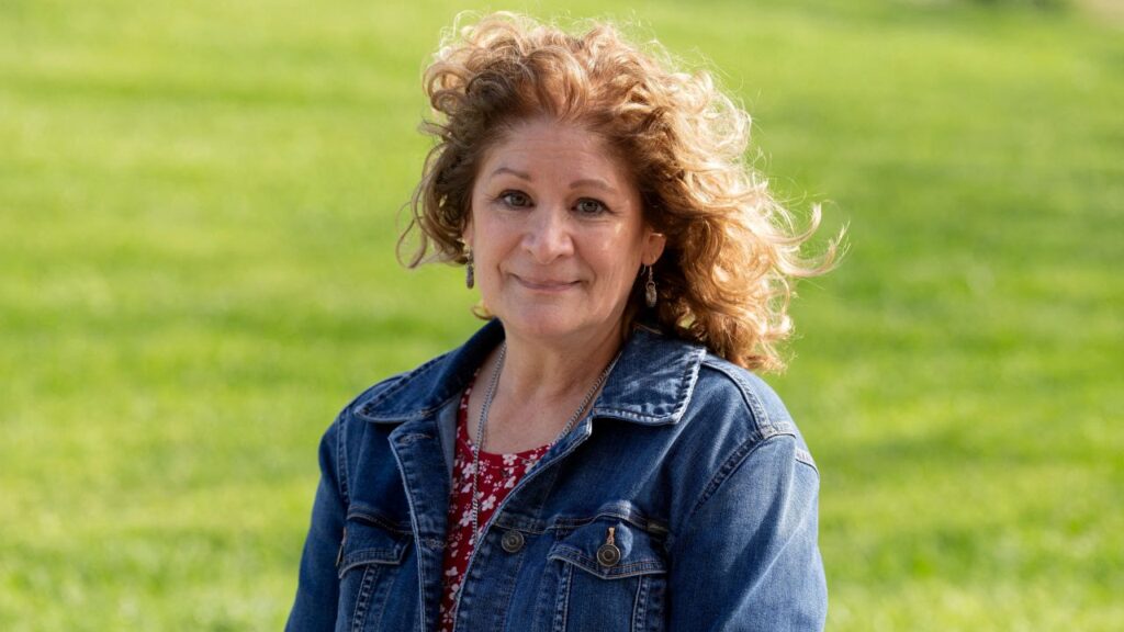 Image of a woman with reddish brown hair wearing a blue jeans jacket standing in a field of green grass