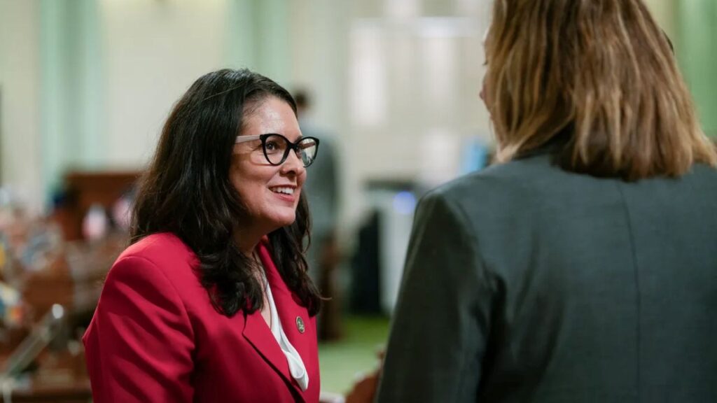Image of a woman facing right and wearing a red dress talking to a woman in a green jacket whose back is to the camera