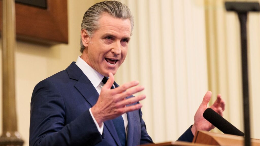 Image of Gavin Newsom in a blue suit, white shirt, and blue tie facing right as he speaks at a podium while gesturing with his hands