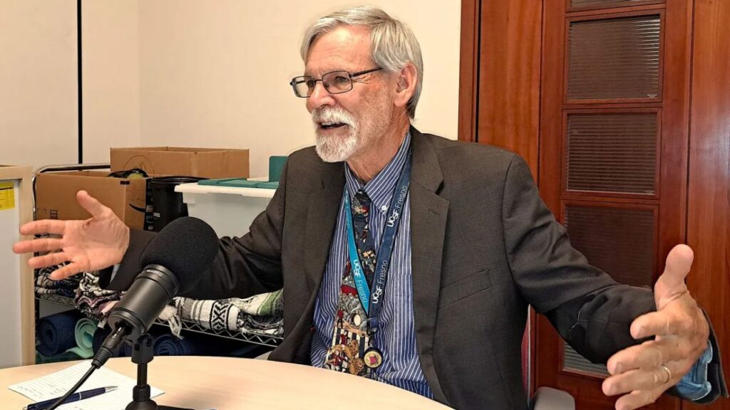 Image of a man in a suit facing left while sitting at a table and talking into his microphone with his arms spread open and his hands turned sideways