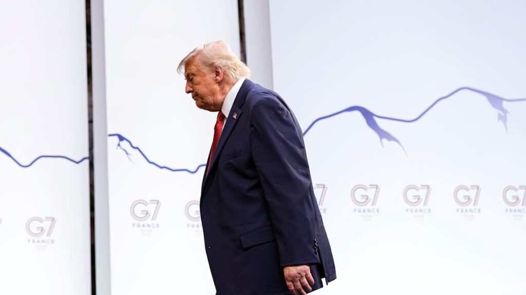 Image of President Trump in a blue suit, white shirt, and tie walking left with his head down in front of a G7 Summit sign