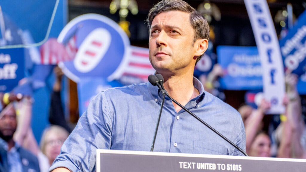 Image of Sen. Jon Ossoff speaking at a campaign rally in a blue open-collar shirt flanked by signs and supporters