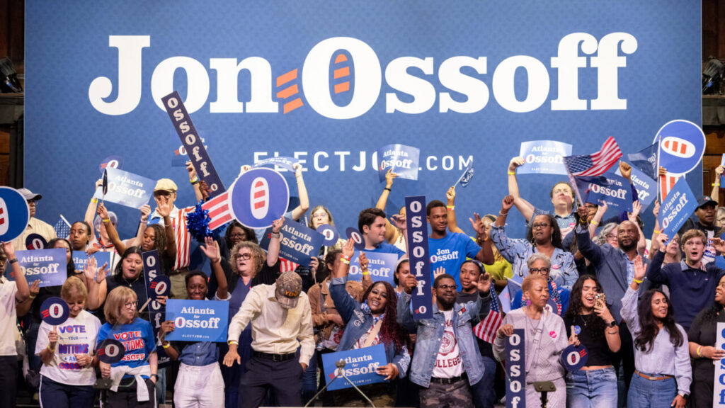 Image of Sen. Jon Ossoff and his supporters on stage at a campaign rally in front of a blue backdrop with his name in white letters