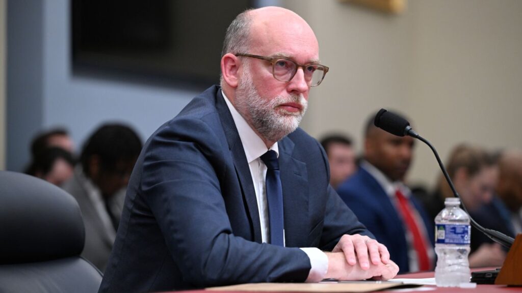 Image of federal budget director Russell Vought, who has a silver beard, looking left while seated at a table and wearing a blue suit.