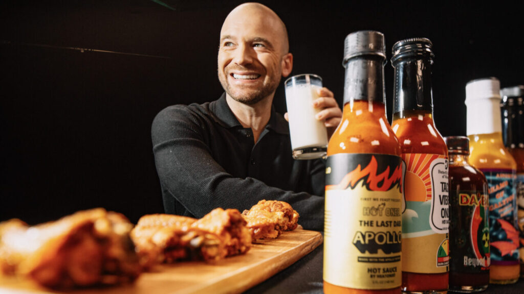 Image of Sean Evans holding a glass of milk, with hot wings sauces and hot wings in the foreground