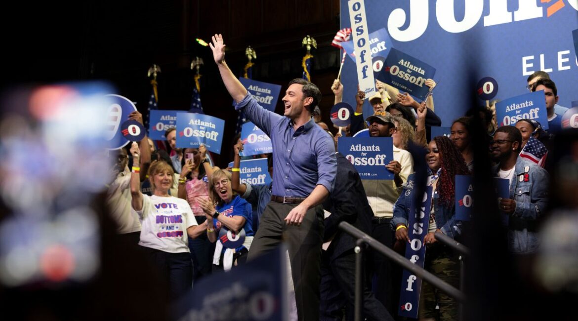 Image of Jon Ossoff facing left and saluting his supporters. He's in a blue open-collar shirt with charcoal slacks