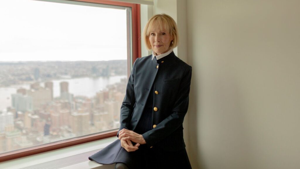 Image of a woman with blond hair standing next to a window. She's wearing a tailored green suit with gold buttons