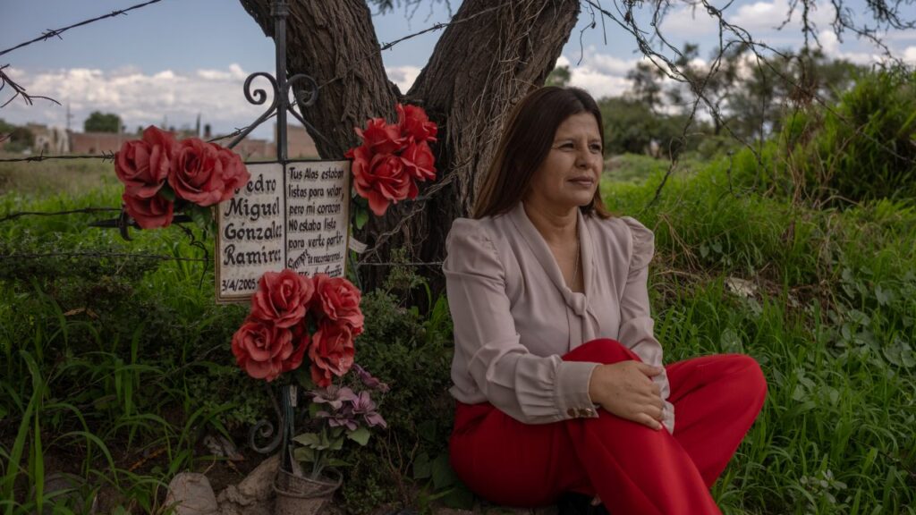Image of a woman in a mauve top and red pants sitting under a tree at a makeshift memorial for a dead student. She is facing right.