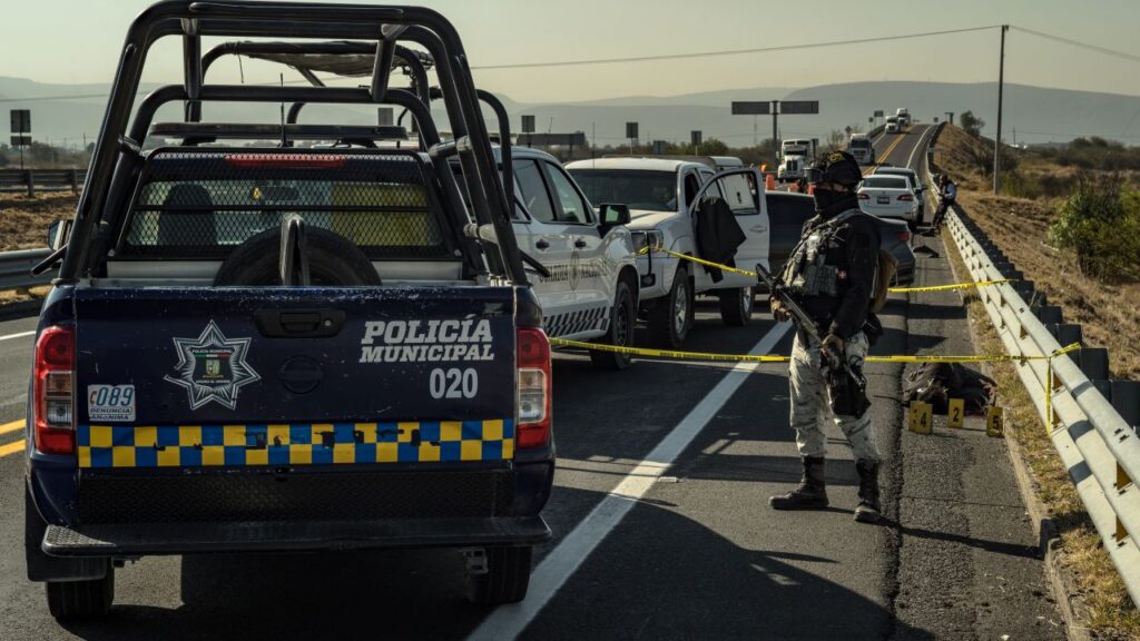 Image of an open bed Mexican police vehicle parked on a highway behind a long row of cars with a police officer armed with a rifle nearby
