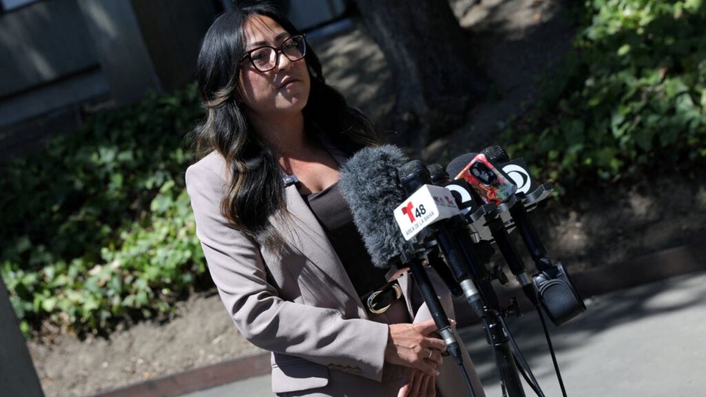 Image of a woman with black hair and glasses facing right and speaking to unseen media