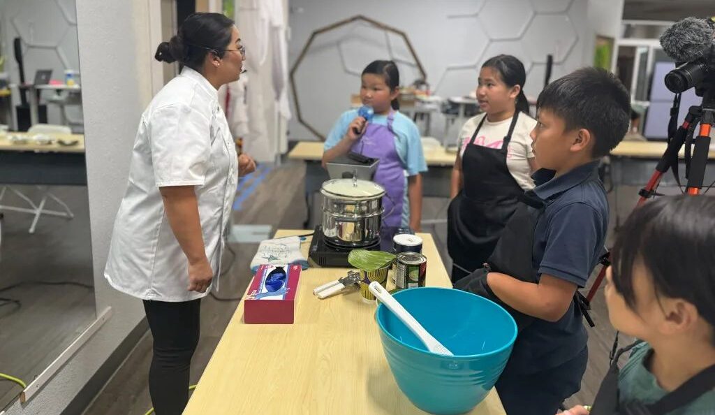 Image of a woman in a white chef's smock facing right and talking to her Hmong cooking students over a kitchen prep table