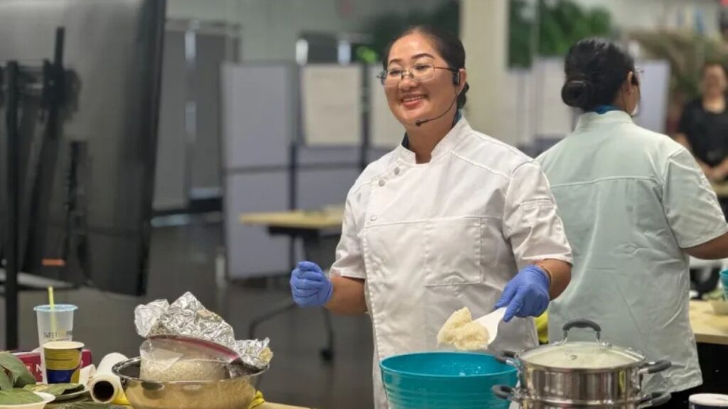 Image of a woman in a chef's smock facing left and teaching a Hmong cooking class