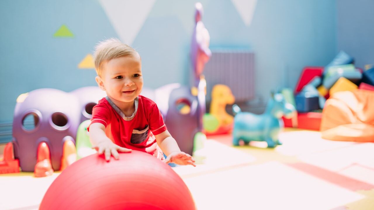 Image of a toddler in a red t-shirt pushing a red ball in a childcare center