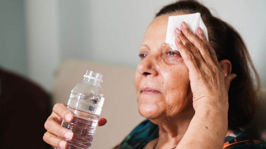 Image of an elderly woman facing left and wiping her forehead and holding a bottle of water while enduring high summer heat