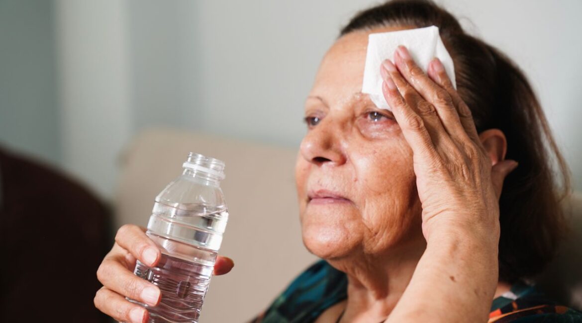 Image of an elderly woman facing left and wiping her forehead and holding a bottle of water while enduring high summer heat