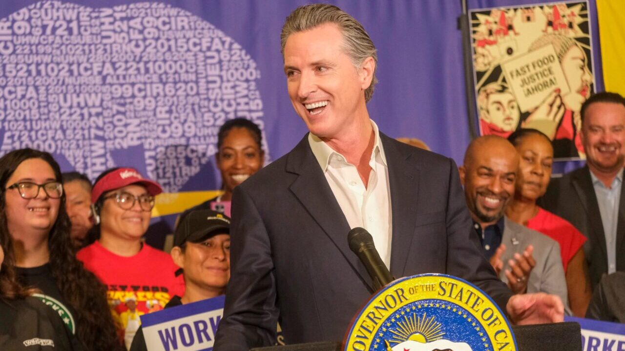 Image of Gov. Newsom looking left. He's wearing a dark blazer and an open collar shirt while surrounded by minimum wage increase supporters