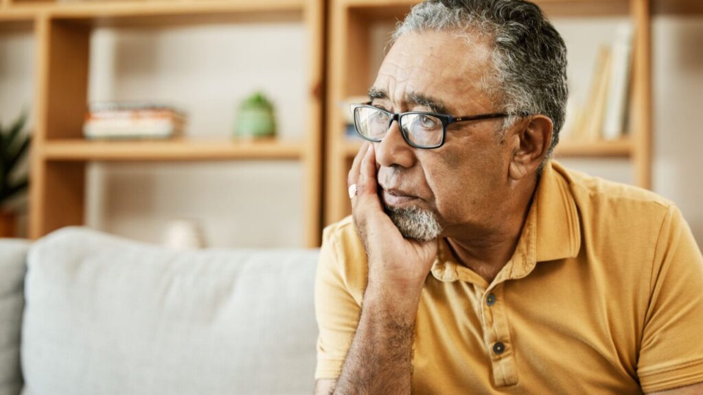 Image of a senior man facing left with his hand on his chin staring into space. He's wearing a tan polo shirt and sitting on a white couch