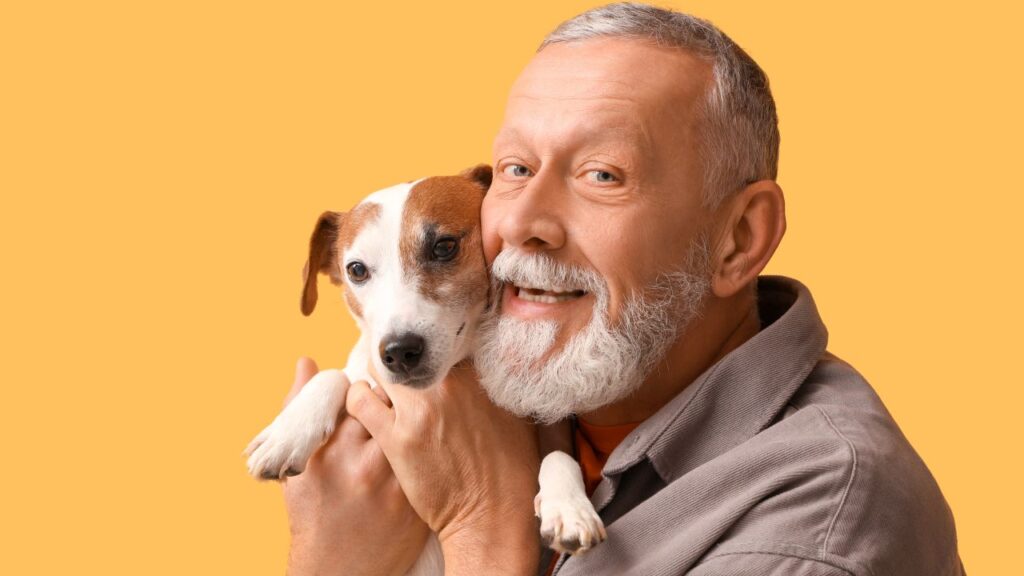 Image of a senior man with a beard facing left and clutching his small mixed breed dog to his face. The image has an orange backdrop.