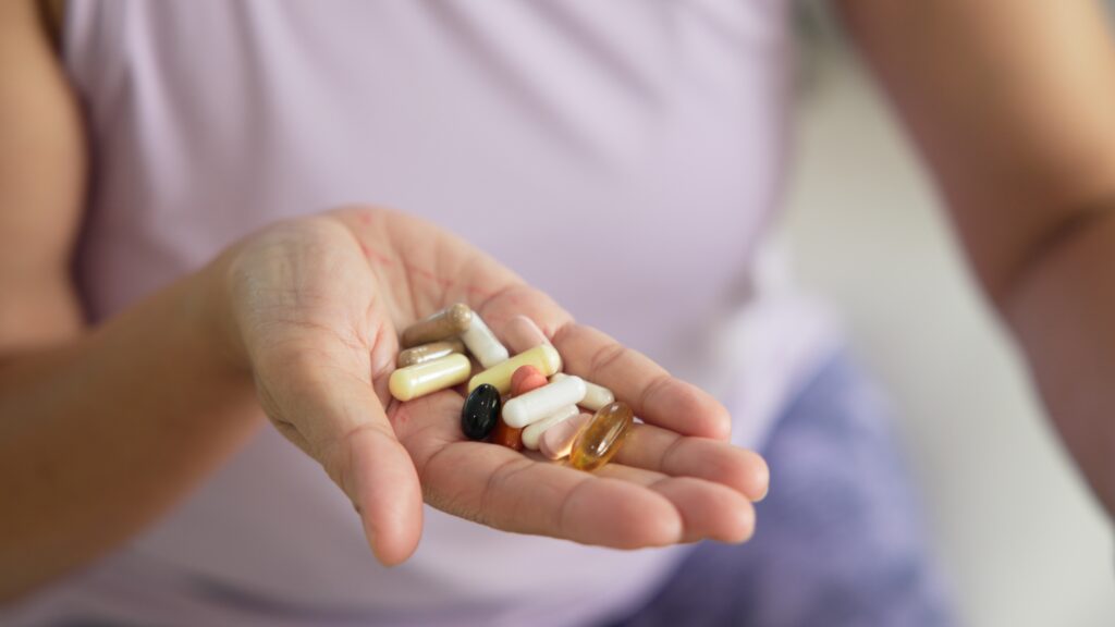 Image of a woman in a lilac blouse with her hand extended and holding many supplements, vitamins, and medications