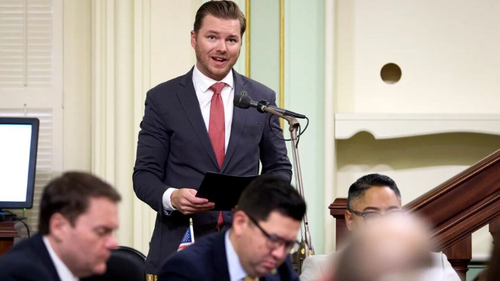 Image of Republican Assemblymember Greg Wallis standing and speaking. He's wearing a dark suit, white shirt, and red tie and has a stubble beard