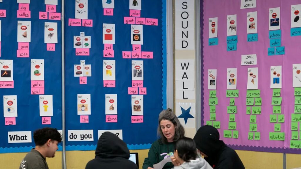 Image of a reading teachers facing left and working with students around a table in a brightly colored classroom with mini-posters of words, letter sounds, and pictures on the wall