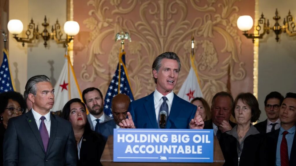 Image of Gov. Gavin Newsom in a dark suit, white shirt, and red tie in front of a blue-and-white Holding Big Oil Accountable banner