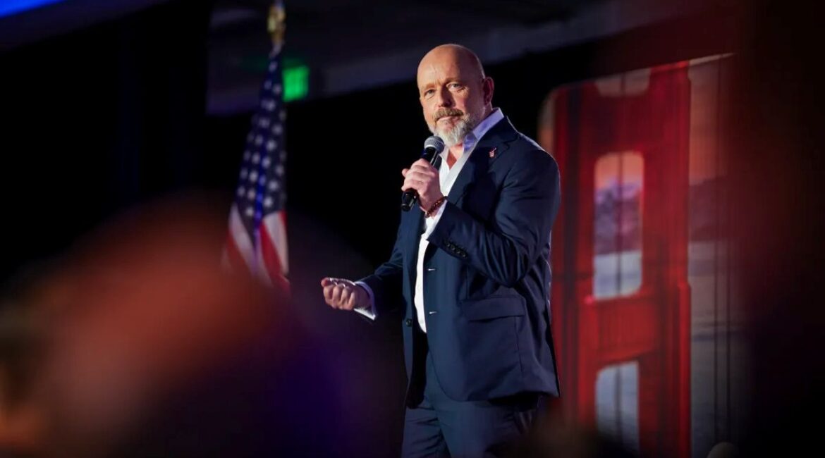 Image of Steve Hilton in a blue suit and white open collar shirt standing on a stage with a microphone in his hand and flanked to the left by the American flag