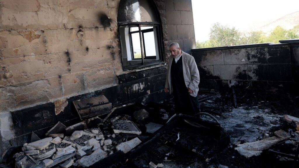 Image of a man in a light colored coat and dark sweater inspecting arson damage in a Palestinian mosque