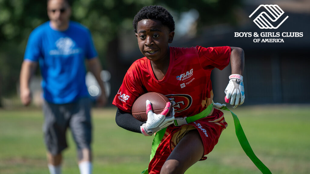 Image of a young flag football player in a red uniform running with the ball as a coach dressed in blue watches
