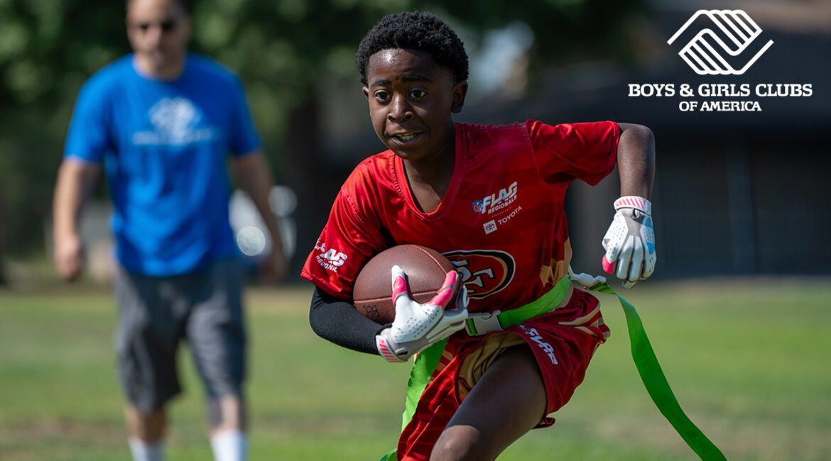 Image of a young flag football player in a red uniform running with the ball as a coach dressed in blue watches