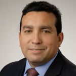 Portrait of David Magaña in a dark suit, blue shirt and red tie