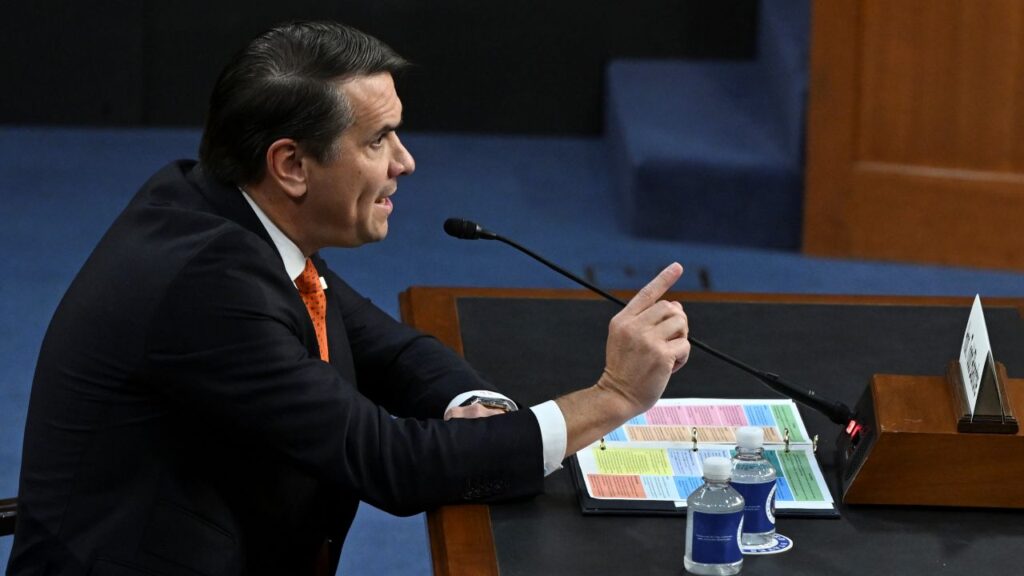 Image of Todd Blanche in a dark suit, white shirt and orange tie facing left and speaking while seated and pointing his index finger