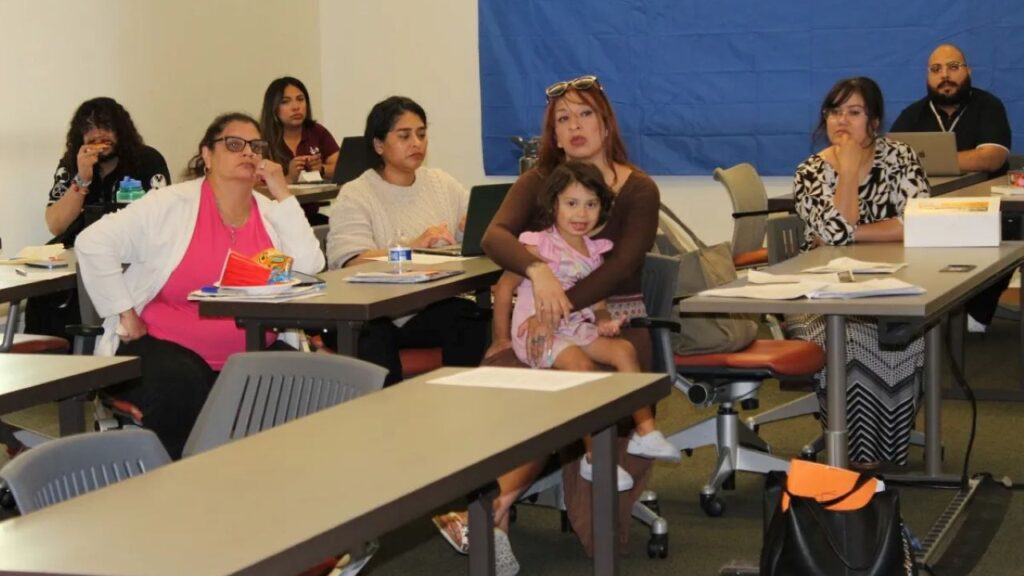 Image of a meeting of mostly women and mothers sitting at tables and listening to an unseen speaker