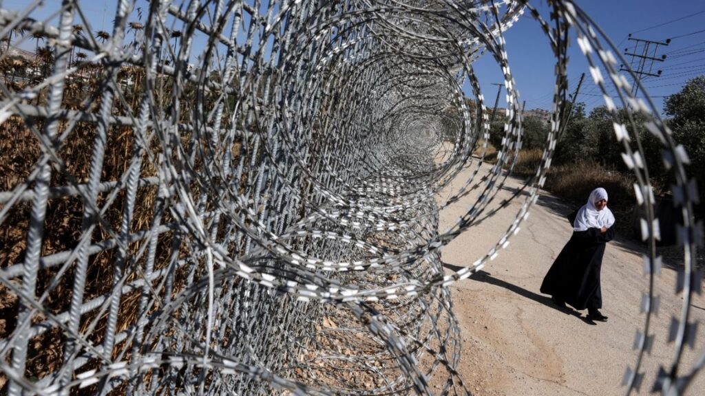 Jawaher Foqahaa walks past the tall metal fencing installed by the Israeli military that closed off four of the five entrances to their town and runs directly past the family home, in Sinjil, near Ramallah, in the Israeli-occupied West Bank, June 26, 2026. (Reuters/Ammar Awad)