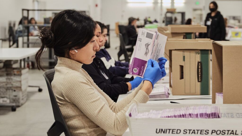 Image of a woman with dark hair and wearing an oatmeal colored top facing right as she sorts through mail-in voting ballots
