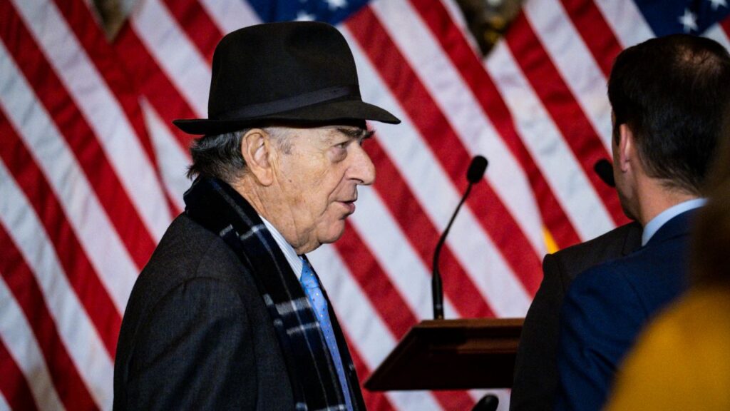 Image of Paul Pelosi wearing a formal hat in dark attire facing right in front of an American flag