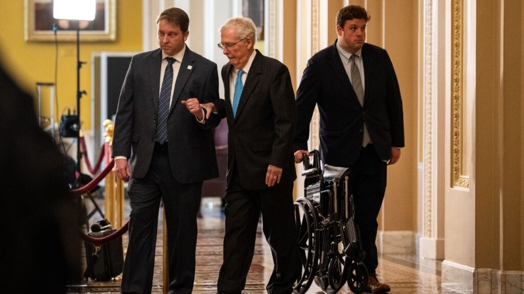 Sen. Mitch McConnell (R-Ky.), aided by two staff members, makes his way to a Senate Republican luncheon on Capitol Hill in Washington, May 12, 2026. McConnell, who has been hospitalized for six weeks, said on Monday, July 27, that he would not be returning to the Senate this week as lawmakers work toward their August recess. (Haiyun Jiang/The New York Times)
