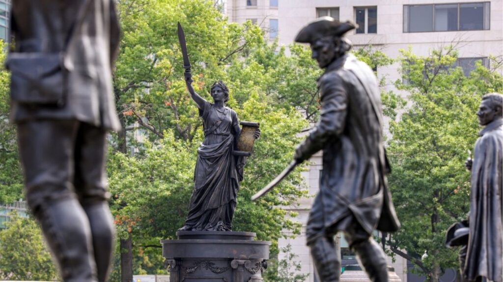 Some of the 21 statues of soldiers and Revolutionary scenes designed by Matthew Taylor, a member of the federal Commission of Fine Arts, are seen at Freedom Plaza in Washington on July 16, 2026. The grouping includes one statue, center left, that Taylor called “a new icon of American liberty.” (Valerie Plesch/The New York Times)