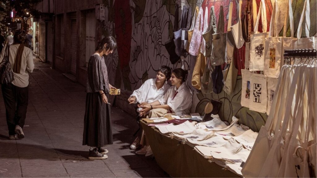 Young Iranians sell tote bags as people walk past on Enghelab street in the center of Iran's capital, Tehran, July 7, 2026. A journey through wartime Iran reveals a nation of many layers: defiant but also weary, despairing but hopeful. (Emile Ducke/The New York Times)