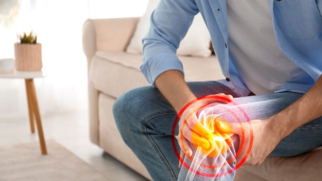 Composite image/illustration of a man in jeans, white t-shirt and a blue shirt sitting and holding his knee, indicating pain with an illustration of the bones and ligaments inside the knee