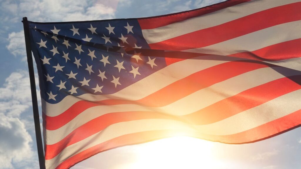 Image of US Flag waving beneath a blue sky and reflecting the sun