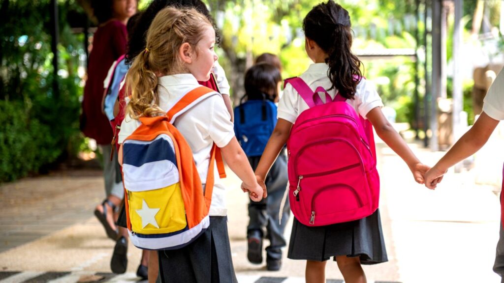 Image of young students holding hands as they walk to school with brightly colored backpacks.