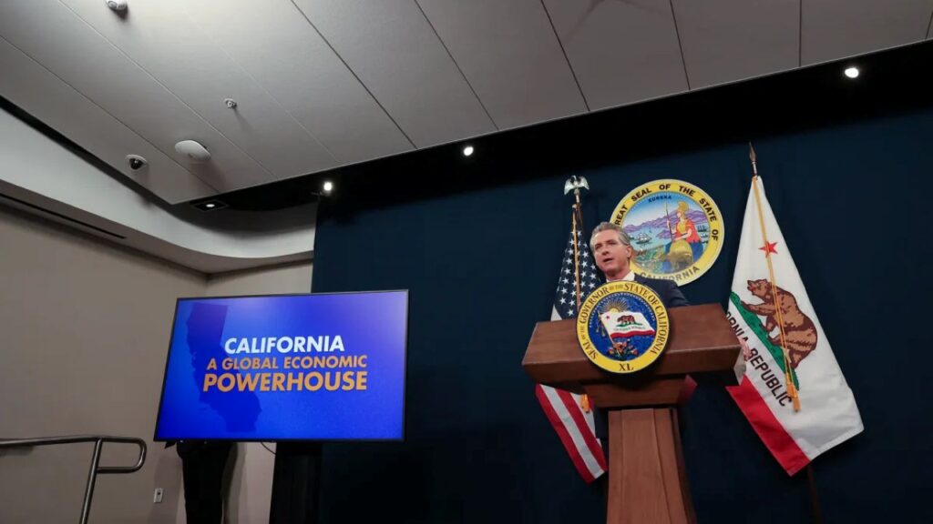 Image of Gavin Newsom facing left while standing at a lectern and leading a news conference about the state budget