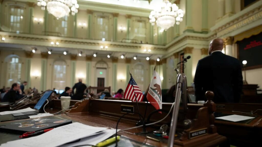 Image of the interior of the California Assembly room with crystal chandeliers, American and California flags, Assemblymembers, and desks