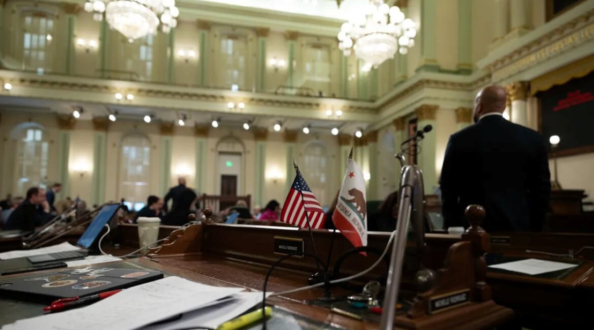 Image of the interior of the California Assembly room with crystal chandeliers, American and California flags, Assemblymembers, and desks