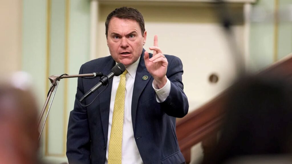 Image of Assemblymember Carl DeMaio in dark suit, white shirt, and yellow tie speaking at a microphone and pointing his index finger up