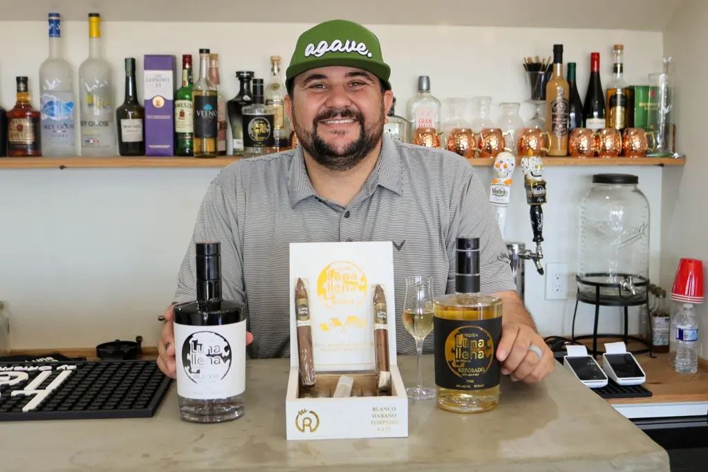 A man in a baseball cap sitting at a table behind his agave liquor and cigar products